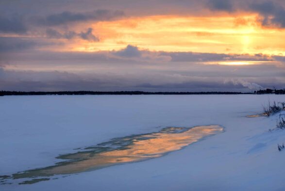 Jäinen luminen joki, tummaa vastarantaa ja keltainen taivas, jota halkovat siniharmaat pilvet.